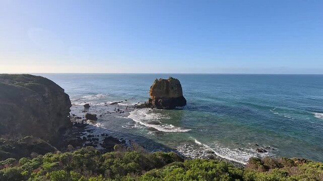 Rocky sea stack rises just offshore from a scrubby headland as gentle waves roll in beneath a bright blue sky.