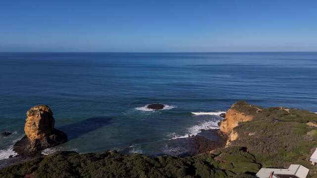 Rock sea stack and low cliffs lead toward a white lighthouse on a calm, sunny coastal headland as the drone glides forward.
