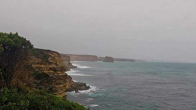 Layered sandstone cliffs drop to choppy ocean swells with misty rock stacks fading into an overcast horizon.
