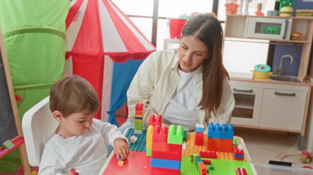 Woman guiding little boy stacking colorful blocks on a table, hand placing a block on a tower in a building; nurturing learning.