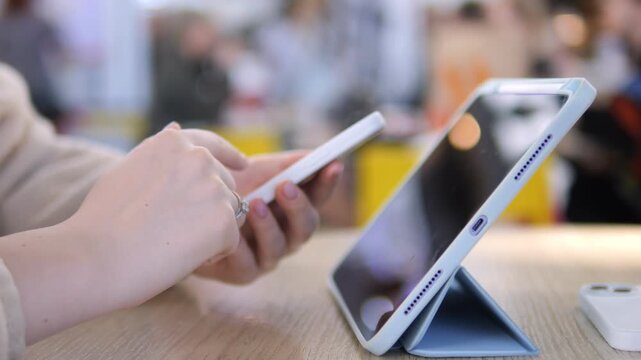 Close-up of hands using a smartphone. Tablet rests upright on table nearby. Person appears focused and calm. Suitable for tech reviews, digital learning, or retail settings