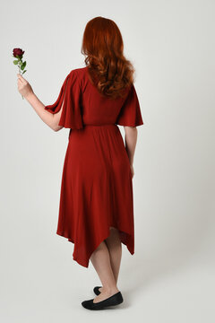 Full length portrait of beautiful redhead model wearing elegant red modern dress. standing pose, walking away facing backwards, holding a romantic red rose, isolated figure studio background