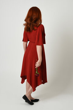 Full length portrait of beautiful redhead model wearing elegant red modern dress. standing pose, walking away facing backwards, holding a romantic red rose, isolated figure studio background