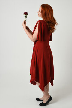 Full length portrait of beautiful redhead model wearing elegant red modern dress. standing pose, walking away facing backwards, holding a romantic red rose, isolated figure studio background