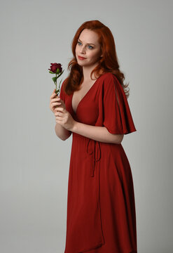 close up portrait of beautiful model with long red hair, wearing an elegant red modern dress. standing pose holding a romantic red rose, isolated figure on studio background.
