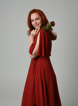 close up portrait of beautiful model with long red hair, wearing an elegant red modern dress. standing pose holding a romantic red rose, isolated figure on studio background.
