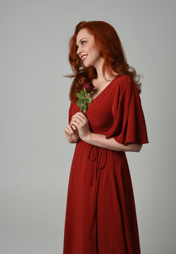 close up portrait of beautiful model with long red hair, wearing an elegant red modern dress. standing pose holding a romantic red rose, isolated figure on studio background.
