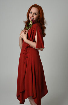 close up portrait of beautiful model with long red hair, wearing an elegant red modern dress. standing pose holding a romantic red rose, isolated figure on studio background.
