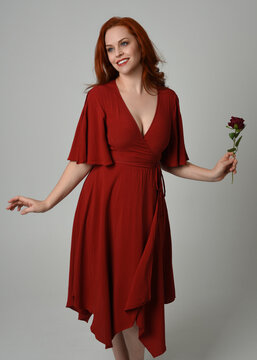 close up portrait of beautiful model with long red hair, wearing an elegant red modern dress. standing pose holding a romantic red rose, isolated figure on studio background.
