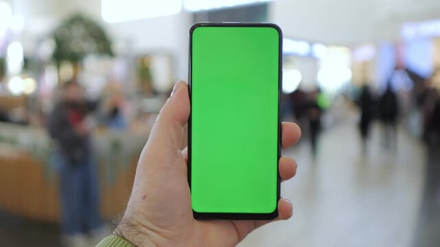 Hand holds phone with green screen in busy indoor space. Blurred background shows people walking casually. Focus on device for app or advertisement display. Suitable for UI, marketing