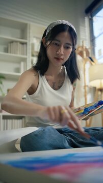 Young Asian woman artist painting on canvas easel with paintbrush in art studio, representing creative professional women designer, fine arts student lifestyle and freelance illustrator