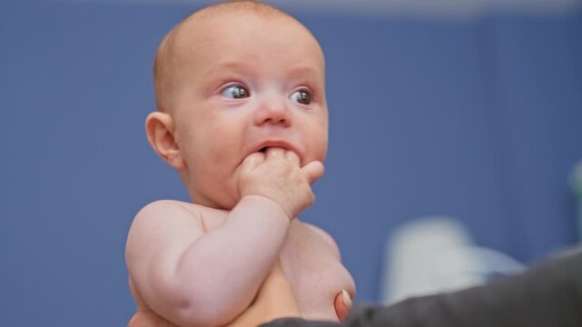 Baby girl with fingers in mouth, caregiver hand supporting bare chest in studio blue backdrop; curiosity bonding.