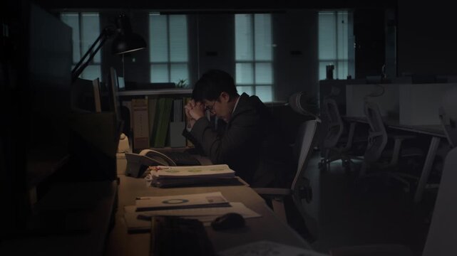A stressed businessman sitting at a classic wooden desk with his head down in a dark office late at night with a warm lamp light.