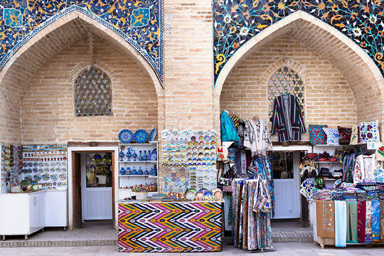 Souvenir shops inside Nadir Divan Begi Madrasah in Bukhara displaying colorful ceramics and textiles beneath ornate tiled arches. Ideal for travel publications and cultural tourism content.