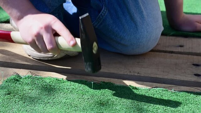 a person's hands using a hammer to carefully drive a small nail into a rough wooden plank, securing a piece of bright green artificial turf.