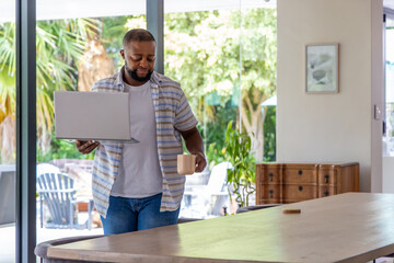 Fototapeta premium African American man standing in dining area holding open laptop and ceramic mug near doors