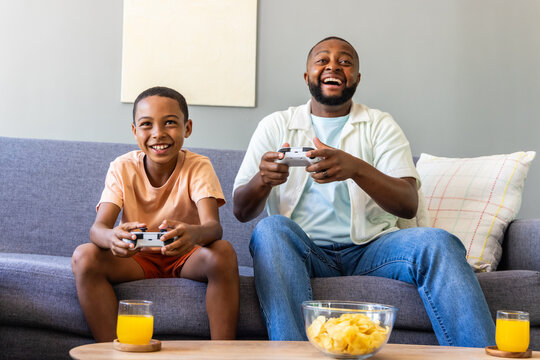African American father, youth male sitting on gray sofa gaming with white pads chips, orange juice