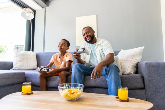 African American dad and teen son sitting on sofa playing with controllers, chips, juice on table