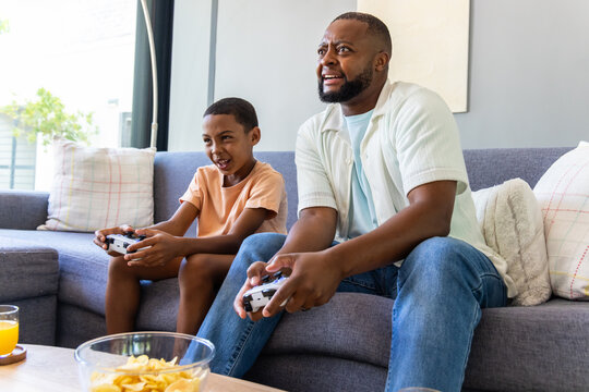 African American father-child sitting on sofa at home holding white gamepads sharing chips and OJ