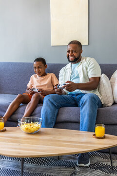 African American father and child playing with controllers on gray sofa at home with orange juice