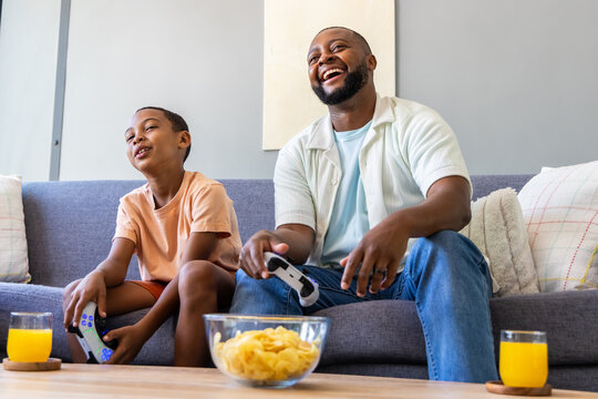 African American father and son sitting on gray couch at home playing games with controllers, chips