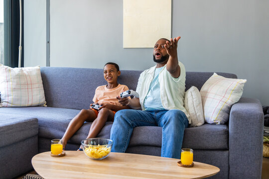 African American father and child playing games, reacting on sofa with controllers, chips, juice