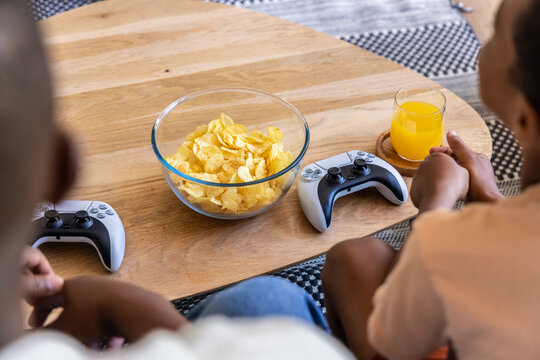 African American dad and son playing with wireless controllers at coffee table with chips and juice