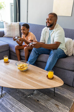 African American father and son sitting on gray sofa playing with white controllers, chips, juice