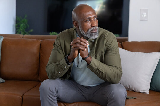 Senior African American man sitting on leather couch at home, hands clasped, eyeglasses on cord