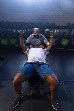 African men, lifter in white tee shorts doing hex dumbbell press on flat bench, spotter spotting