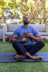 Obraz premium African American man sitting on yoga mat in backyard, with hands over chest near wooden sofa