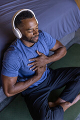 Fototapeta premium African American man meditating on green mat beside bed, wearing blue shirt and white headphones