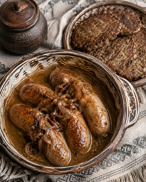 Traditional Belarusian homemade pork sausages verashchaka served with fried onions in gravy sauce and potato pancakes draniki in ceramic bowls on a rustic tablecloth.