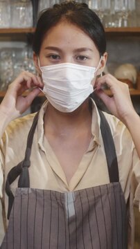 Vertical Screen : Young Asia female restaurant staff wearing protective face mask using infrared thermometer checker temperature gun on customer's forehead before enter. Lifestyle after corona virus.