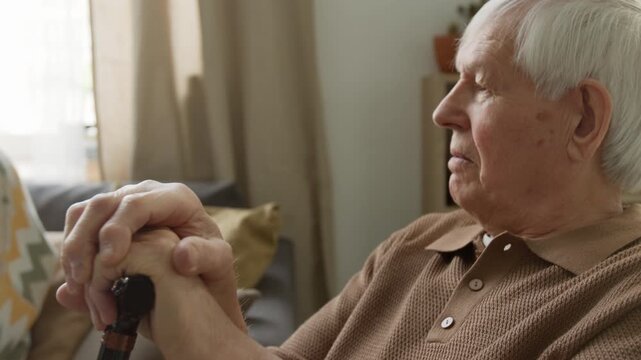 Side view shot of senior man sitting on sofa and holding walking cane with both hands while senior woman sits in background. Video for senior care services and retirement home promotions