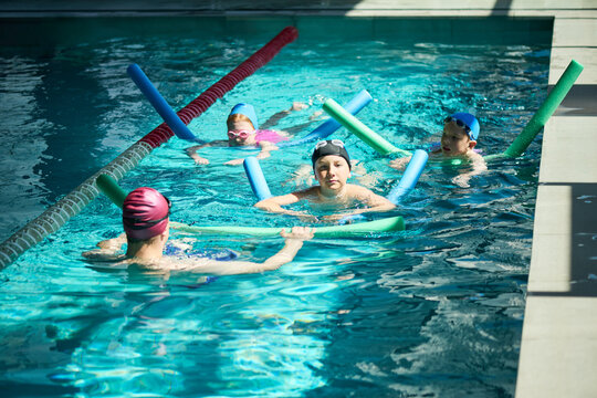 Wide shot of children practicing swimming with instructor in pool, using foam noodles during lesson, building water safety and fitness. Useful for youth sports, swim school promotion