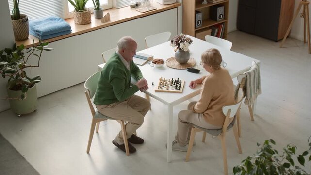 Wide angle shot of senior man and senior woman playing chess at table in retirement home. Clip for marketing campaigns promoting active aging and senior care facilities