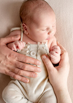 Baby sleeps in beige romper on soft blanket. Adult hands provide comforting support.