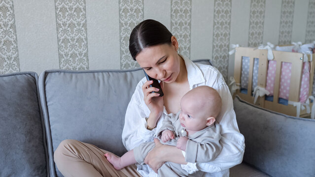 Young mother multitasking on a sofa, holding her newborn while talking on a smartphone modern parenting, work from home juggling, caring, connection and everyday stress at home