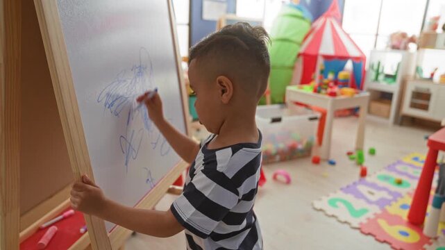 Little boy drawing on a whiteboard easel with marker in a bright playroom studio surrounded by toys and foam numbers; creativity concentration.