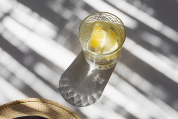 Water glass with sparkling bubbles, summer refreshing lemon drink with straw hat, caustic sunlight shadows on aesthetic background. Holiday and vacation concept. © juliasudnitskaya