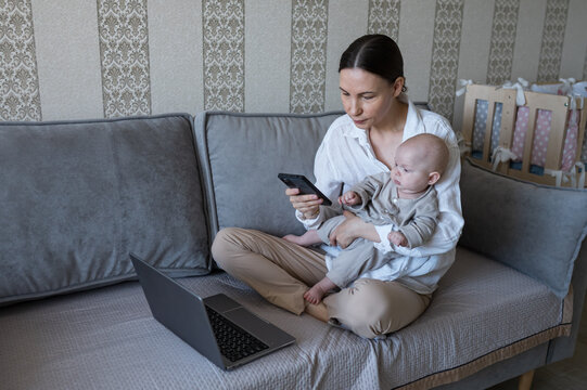 Young mother balancing motherhood and career, holding her baby while using a smartphone and laptop on a sofa, embodying remote work and modern parenting challenges from home
