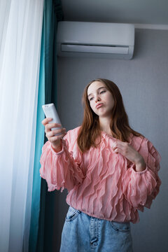 Young frustrated woman holding a remote control, operating the air conditioning unit indoors, seeking relief from hot temperature and feeling uncomfortable during summer