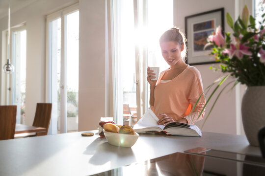 Young woman in kitchen having breakfast in kitchen, paging through book