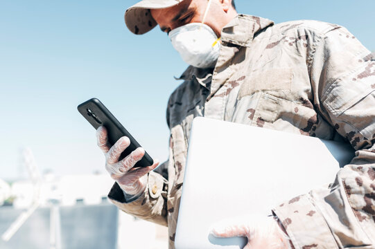 Soldier with face mask on emergency operation, using smartphone, carrying laptop