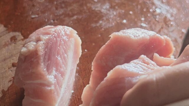 A high-quality, macro close-up video capturing a person precisely slicing fresh raw pork loin meat with a sharp stainless steel knife on a rustic wooden cutting board