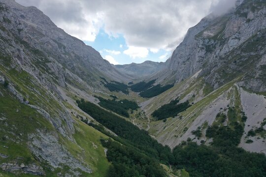 Aerial view of Prati di Tivo valley with rocky mountain slopes, lush green forests, and a winding path under a cloudy sky in Prati di Tivo, Abruzzo, Italy.