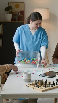 Young female nurse hosting bingo game for seniors in retirement home, holding colorful bingo ball. Footage promoting senior care services, vertical