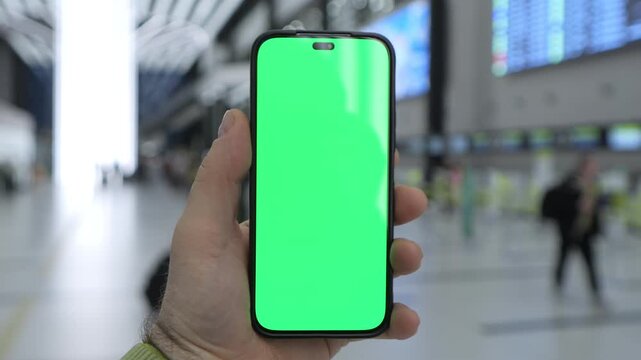 Hand holds phone with green screen. Airport terminal blurred in background. Traveler waits near departure board. Phone shows bright green mockup. People move around with luggage