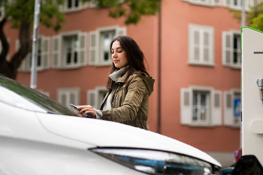 Confident beautiful woman using smart phone for renting electric car at charging station